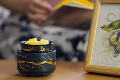Black and gold electric wax melt burner with yellow wax cubes on a wooden table beside a framed bird illustration.