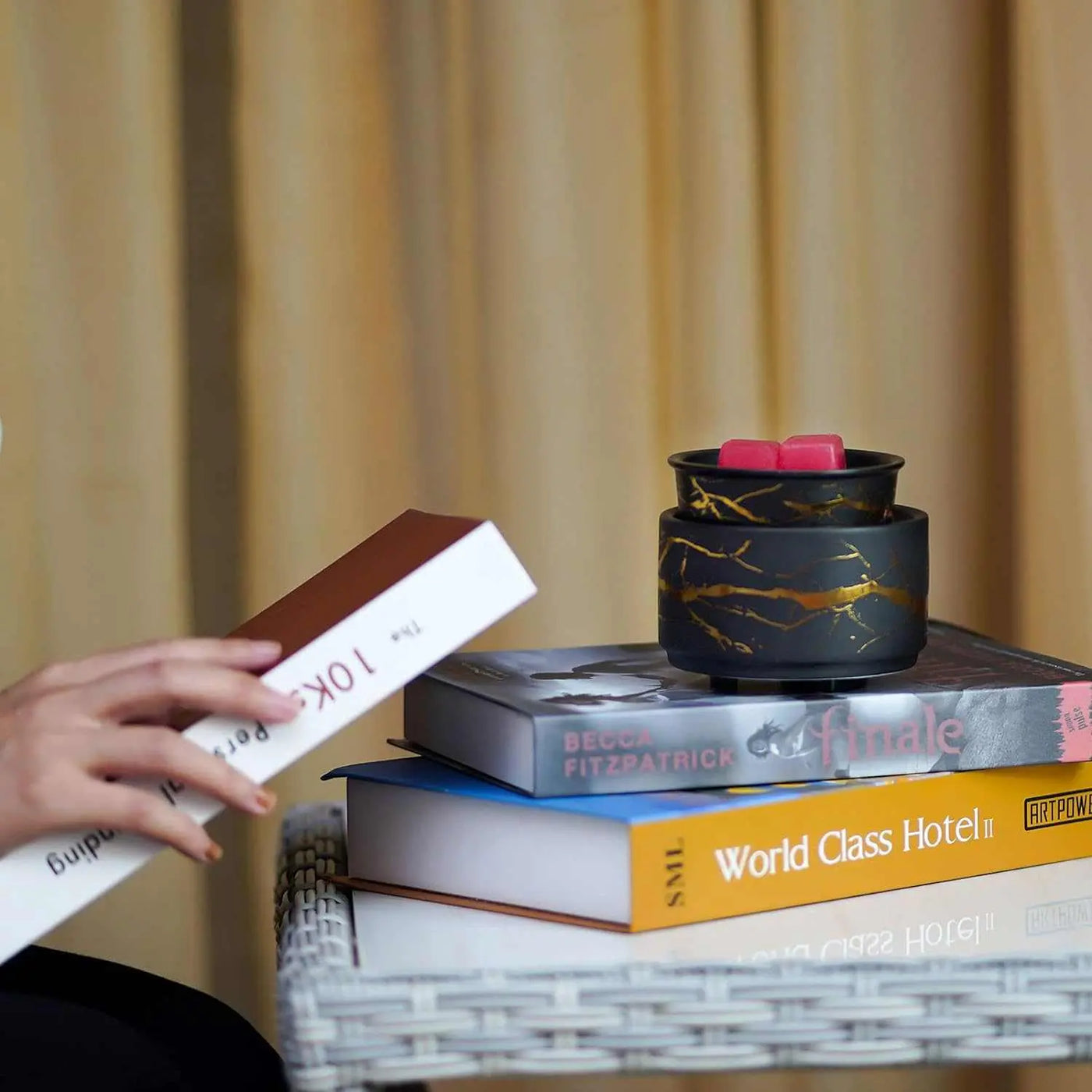 Black and gold electric wax melt burner with pink wax cubes, placed on stacked books beside a person reading.